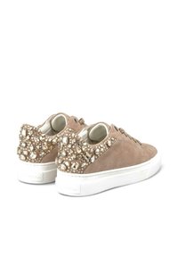 Beige Wildleder-Sneaker mit einer weißen Sohle, die an der Rückseite mit verschiedenen klaren und goldenen Akzenten verziert sind. Die Schnürsenkel vorne verstärken das Design.