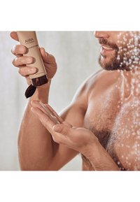 Detergente esfoliante in tubo beige con cappuccio marrone scuro, tenuto sopra un palmo aperto. Un gel trasparente viene erogato, con gocce d'acqua sullo sfondo.