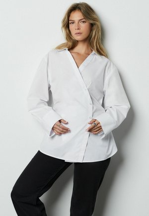 Weißes Langarmshirt mit V-Ausschnitt, Knöpfen vorne und breiten Bündchen. Glatte Textur und maßgeschneiderte Passform. Kombiniert mit schwarzen Hosen.