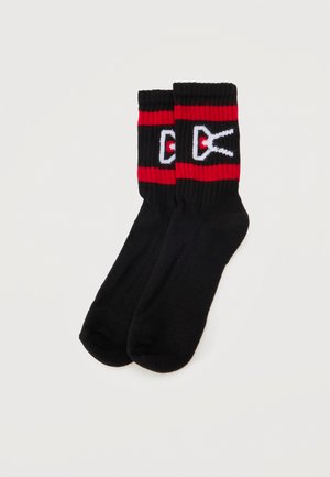Une paire de chaussettes noires mi-mollet, avec des rayures rouges aux bords-côtes et un logo blanc et rouge, sur un fond uni clair.