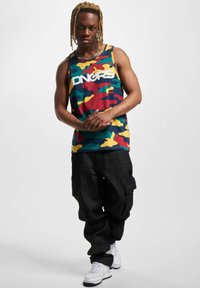 Top de burtă cu camuflaj multicolor și logo-ul "DNGRS", purtat cu pantaloni cargo negri lărgi și teniși albi, evidențiind un stil casual de streetwear.