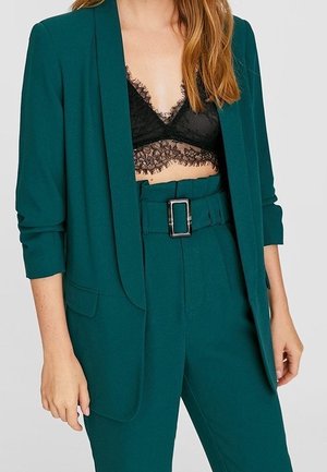 Kvinde iført teal blazer og højttaljede bukser med sort blonde-bralette, hænder afslappet ved siden, mod ensfarvet baggrund.
