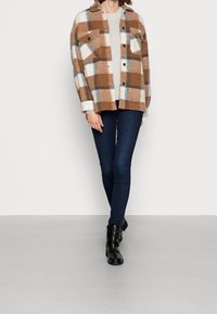 Bruine en witte geruite flanellen jas met twee borstzakken, gecombineerd met donkere skinny jeans en zwarte enkellaarsjes. Effen textuur, ontspannen pasvorm.