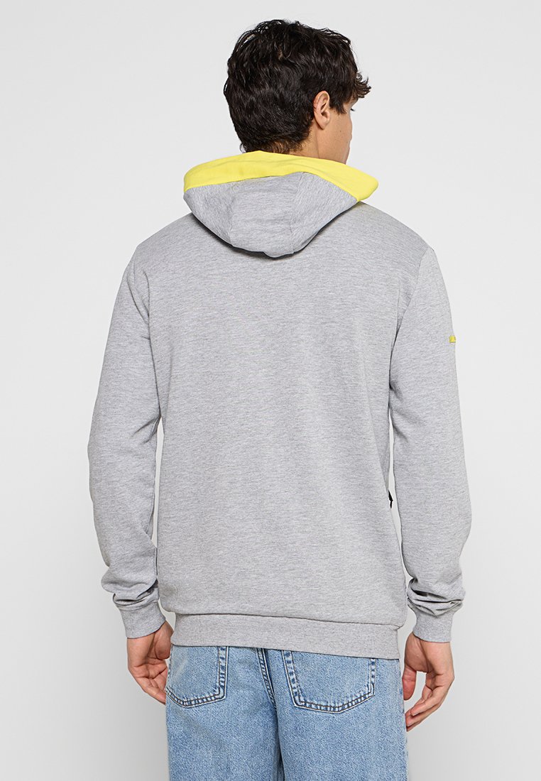 Homme portant un hoodie gris clair avec une capuche intérieure jaune et un jean bleu, se tenant dos à un fond blanc.