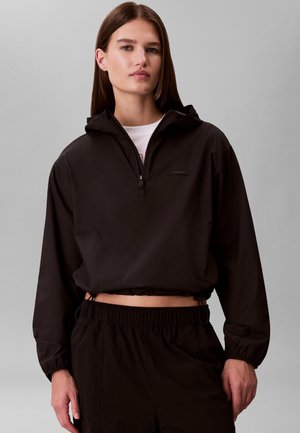 Frau trägt eine schwarze, kurze Kapuzenjacke mit Frontreißverschluss und passende schwarze Hose vor einem einfarbigen grauen Hintergrund.