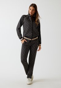 Tuta nero a fantasia con giacca con zip e pantaloni coordinati. Dettagli in oro, polsini a righe e tasche laterali caratterizzano il design.