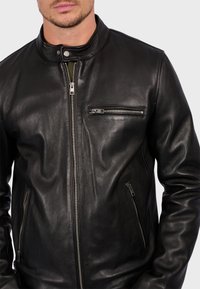 Chaqueta de cuero negro con cuello alzado, cierre frontal de cremallera y bolsillos laterales con cremalleras. Textura suave y diseño ajustado.