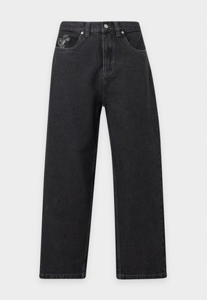Personne portant un jean noir coupe droite, des chaussures noires épaisses et un haut à manches longues noir à motifs, debout devant un fond uni.