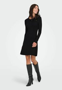 Schwarzes geripptes Strickkleid mit langen Ärmeln, tailliertem Oberteil und schwingendem Rock, kombiniert mit knielangen schwarzen Lederstiefeln mit spitzem Zehen.