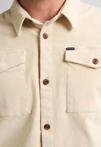 Cămașă din corduroy crem, cu închidere butonată, două buzunare pe piept și butoane maro, având o etichetă mică neagră lângă buzunarul din dreapta.