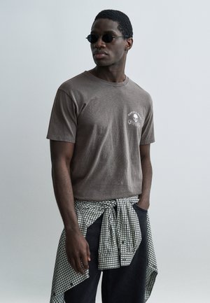 Νεαρός άνδρας που φοράει στρογγυλά μαύρα γυαλιά ηλίου, καφέ μπλουζάκι με σχέδιο και πουκάμισο καρό δεμένο γύρω από τη μέση του, με φόντο ένα μονόχρωμο τοπίο.