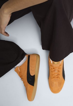 Persona con sneaker in camoscio color cuoio con lati neri e suole color gomma, abbinati a pantaloni neri e calzini bianchi, seduta a gambe incrociate.