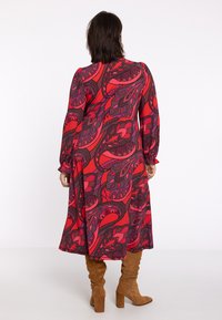 Rotes und lila gemustertes Kleid mit langen Ärmeln, gerafften Manschetten und einem fließenden Saum, getragen mit braunen Wildleder-Stiefeln bis zum Knie.