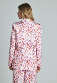 Il blazer floreale presenta un tessuto bianco con fantasie di foglie rosa e viola, un design strutturato, maniche lunghe e una vestibilità sartoriale.