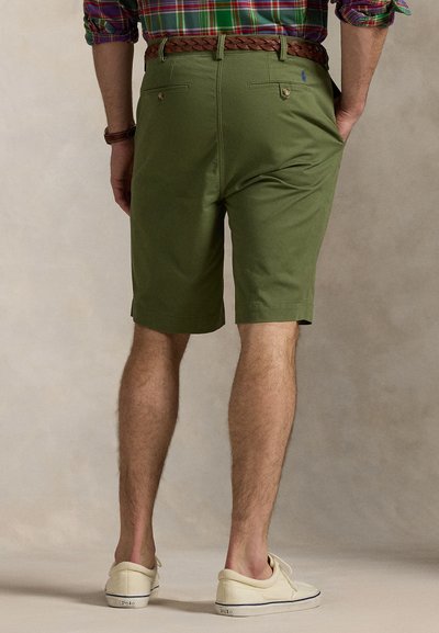 Calções verdes com corte afinado, apresentam bolsos traseiros, um cinto castanho tecido e são combinados com ténis de cor creme.