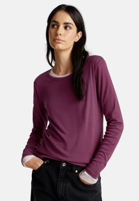 Femme aux longs cheveux noirs portant un t-shirt à manches longues violet superposé sur un haut blanc, les mains dans les poches de jeans noirs, regardant sur le côté.