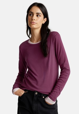 Femme aux longs cheveux noirs portant un t-shirt à manches longues violet superposé sur un haut blanc, les mains dans les poches de jeans noirs, regardant sur le côté.