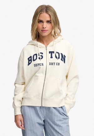 Femme blonde portant un sweat à capuche crème zippé avec le texte "Boston Super Dry Co" et un pantalon bleu clair à fines rayures, debout les mains dans les poches.