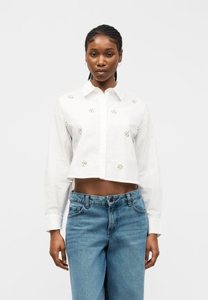 Μαύρη γυναίκα με κοτσίδες που φοράει ένα λευκό κοντό πουκάμισο με μικρές λουλουδένιες λεπτομέρειες και ψηλόμεσα μπλε τζιν, όρθια.