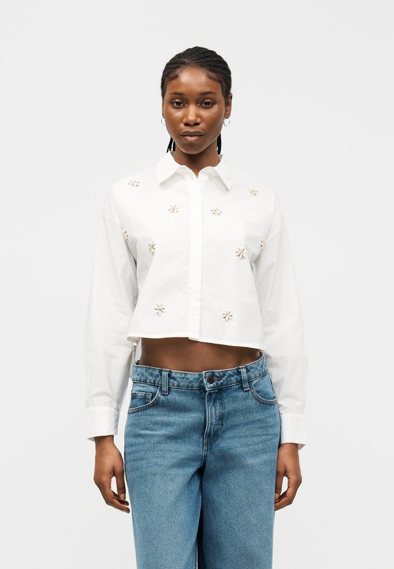 Sort kvinne med flettet hår iført en kort hvit skjorte med små blomsterdetaljer og høytlivs blå jeans, stående rett.