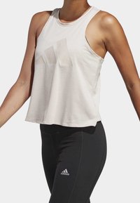 Top atletico senza maniche beige chiaro con un design del logo semi-trasparente, abbinato a leggings neri aderenti con un logo bianco.