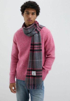 Sciarpa grigia con un motivo a scacchi rosa e nero, drappeggiata attorno al collo, abbinata a un maglione rosa peloso e jeans blu.
