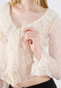Kremfarget bluse i blonder med volanghals og klokkelignende ermer. Stoffer har blomster mønstre som gir et teksturert og delikat utseende.