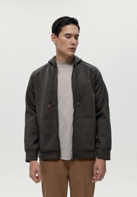 Sivá mikina na zips s hladkým povrchom, nastaviteľnými šnúrkami a prednými vreckami, nosená cez svetlý tričko a hnedé nohavice.