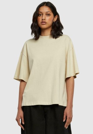 Beige oversized t-skjorte laget av mykt stoff, med rund hals, korte ermer og en avslappet passform, kombinert med mørke bukser.