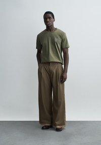 Homme debout, les mains dans les poches, portant un t-shirt vert olive et un large pantalon marron sur un fond clair uni.