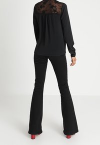 Femme portant un haut noir à manches longues avec un panneau arrière en dentelle florale transparente, un pantalon noir ajusté et évasé, et des chaussures à talons rouges, debout de dos.