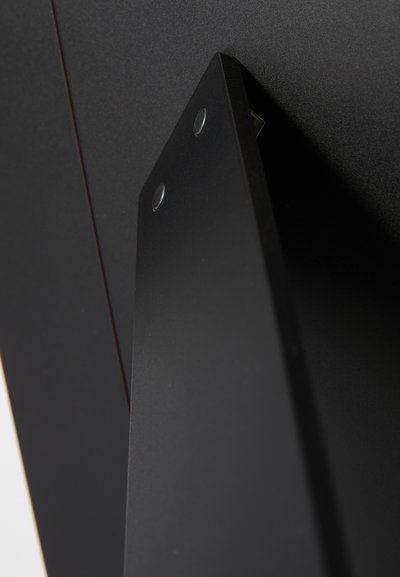 Melns metāla kronšteins ar divām apaļām caurumiem stiprināšanai, ar gludu tekstūru un leņķisku dizainu, piestiprināts pie melnas virsmas.