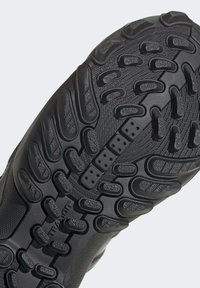 Semelle de chaussure en caoutchouc noir avec une surface texturée, présentant de profonds sillons et des motifs circulaires pour l'adhérence, et un détail de logo au centre.