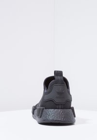 Schwarzer Sportschuh mit strukturiertem Strick-Obermaterial, Gummi-Fersenbereich und markanter gepolsterter Sohle. Verfügt über eine Zuglasche an der Rückseite.