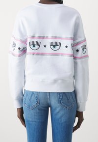Vit sweatshirt med en glittrande rosa och silverrand med tryckta ögon, ögonfransar och stjärnor; matchad med blå jeans med diamantmönster.