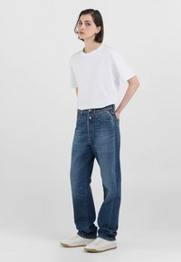 T-shirt bianca a maniche corte abbinata a jeans blu a vestibilità ampia. I jeans presentano una texture sbiadita e un design classico a cinque tasche, abbinati a sneakers bianche.