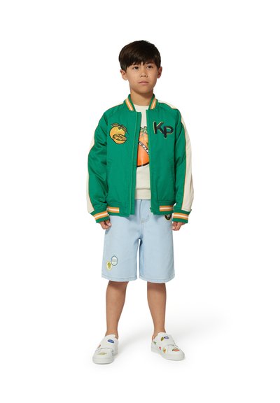 Groene bomberjack met oranje en gele accenten, logo en fruitige patches, gedragen over een licht shirt en blauwe shorts, gecombineerd met bedrukte instappers.