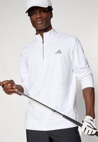 Biele golfové polo s dlhým rukávom a polovičným zipsom, má šedé logo Adidas, hladký povrch, nosené s bielou čiapkou a golfovou rukavicou, drží palicu.