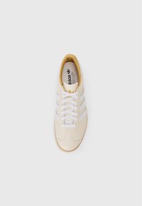 Witte leren sneaker met drie horizontale witte strepen, ronde neus, tan kleurige zool en witte veters. Binnenin zijn merken en details te zien.