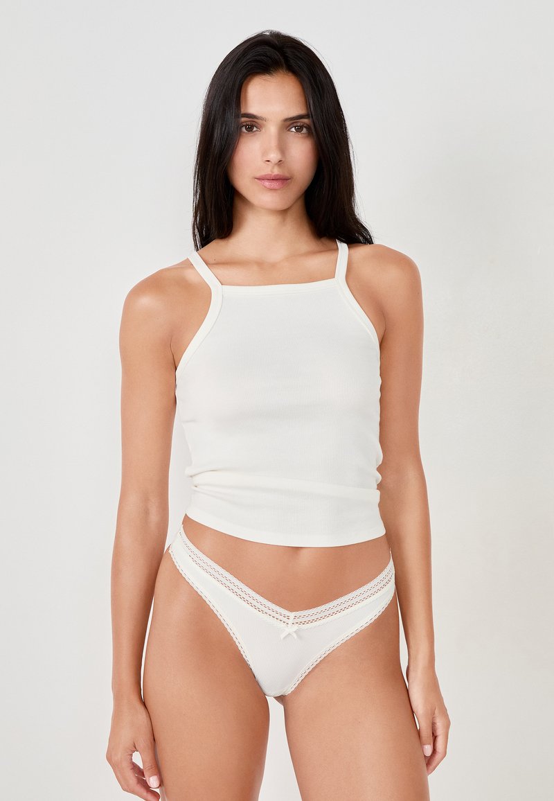 Top a costine bianco senza maniche con spalline sottili abbinato a un perizoma bianco in stile bikini con dettagli in pizzo e un piccolo fiocco sul davanti.