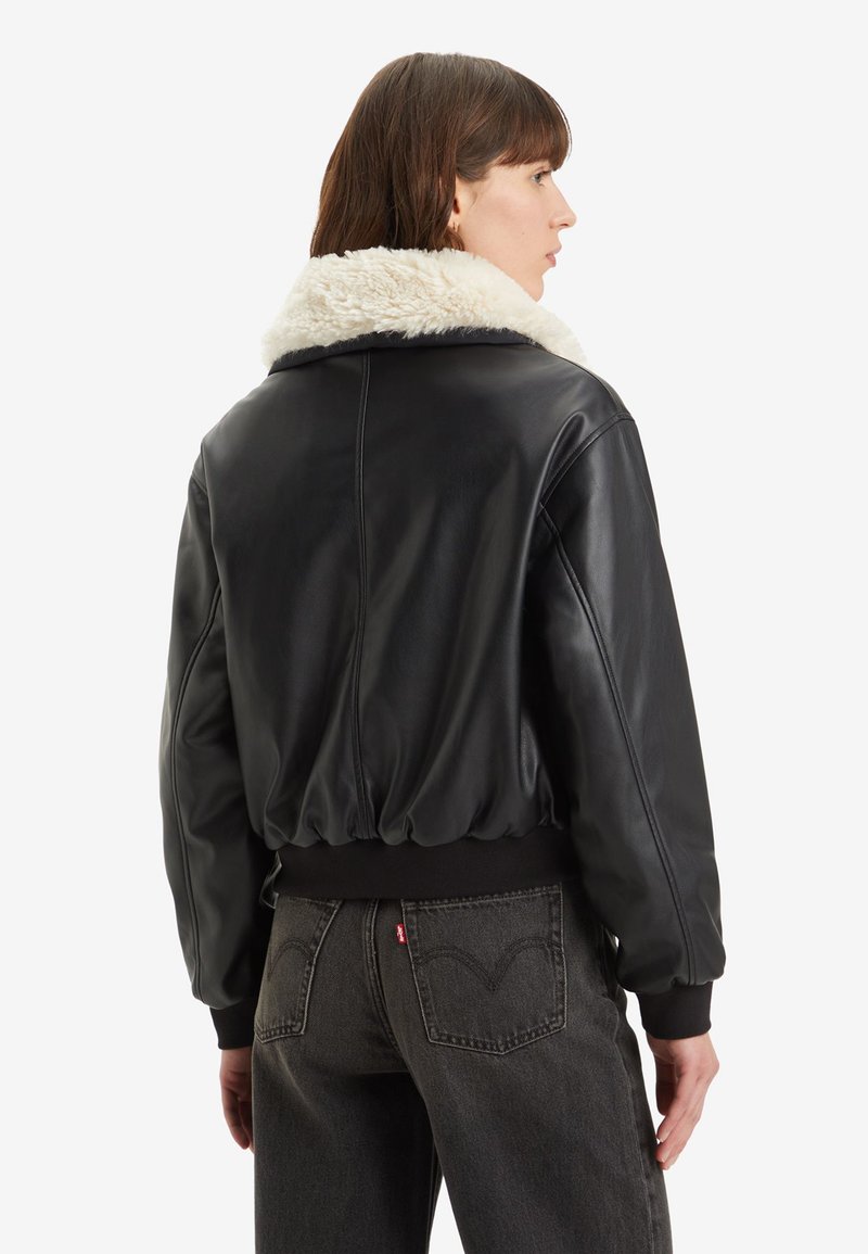 Chaqueta de cuero negra con un collar de piel sintética, cintura ajustada y puños acanalados. Textura suave y diseño redondeado. Se muestra la vista trasera.