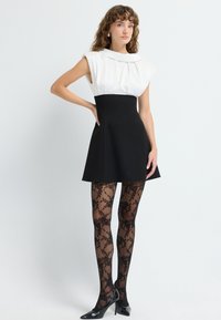 Zwart skateboard jurk met witte gestructureerde lijfje en geplooid detail. Doorzichtige zwarte panty met bloemenpatroon. Zwarte glanzende hakken.