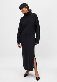 Abito lungo nero in maglia con alto colletto a imbuto, spacco laterale e polsini a coste. Tessuto liscio e silhouette aderente fino alle caviglie.