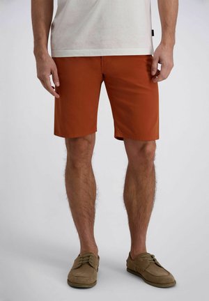 Homme portant un short rouille arrivant au genou, des chaussures décontractées beiges et un t-shirt blanc, debout devant un fond uni.