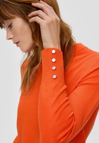 Oranges Sweatshirt mit hohem Kragen und silbernen Knopfakzenten an den Bündchen. Glatte Textur mit figurbetontem Design.