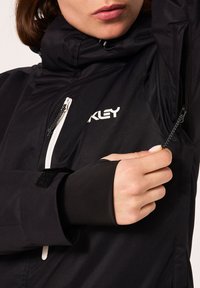 Veste noire imperméable avec col haut, dotée d'un logo blanc, de poches zippées et de poignets avec trou pour le pouce pour une protection supplémentaire.