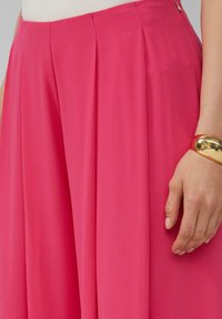 Rosafarbene, plissierte weit geschnittene Hosen aus glattem Stoff. Mit einem seitlichen Reißverschluss und einer Hand einer Person, die ein goldenes Armband trägt, das auf dem Taillenbund liegt.