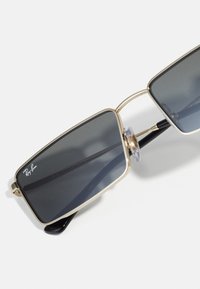 Kaldkaitsega kandilised prillid tumedate hallide objektiividega, kuldse metallraami, läbipaistvate ninalappidega ja mustade atsetaattempelidega, millel on Ray-Ban logo.