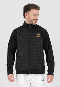 Sort zip-up jakke med et tekstureret mønster, høj krave og et broderet gult logo på brystet, med lange ærmer og ribbede manchetter.