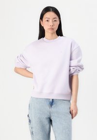 Tânără purtând un hanorac oversized de culoare violet deschis și blugi decolorati, stând cu o mână pe șold pe un fundal alb.